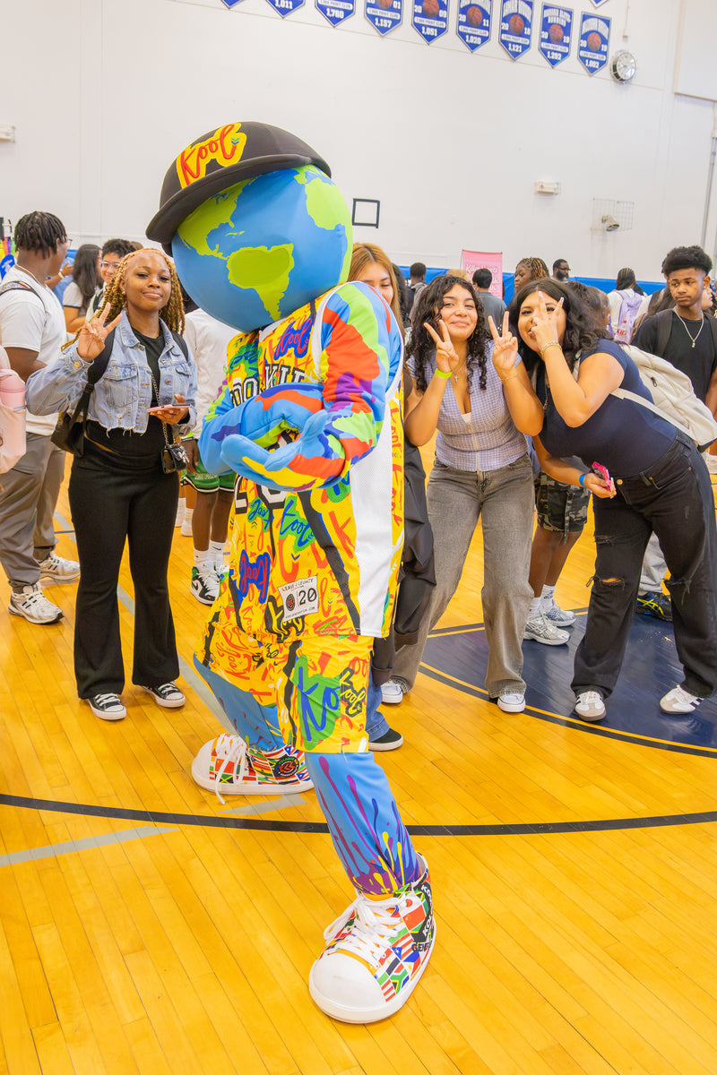 kooli in a tie-dye costume stands on a gymnasium floor, surrounded by students and faculty in a school gym.