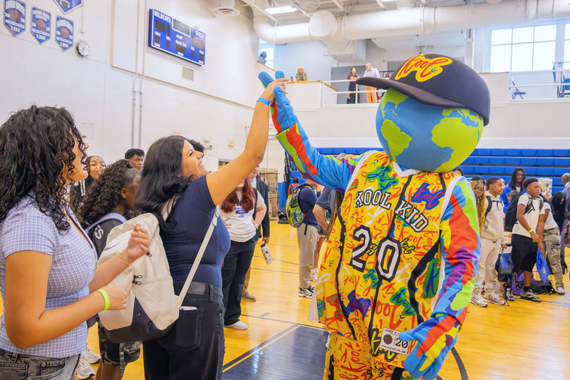 kooli in  basketball jersey and blue cap raises its hand in a gymnasium, with students and faculty standing nearby.