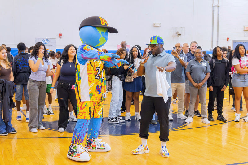 kooli in a basketball uniform with a black cap stands on a gymnasium floor facing a crowd of people, including a man in a light blue polo shirt and black pants holding a white towel.