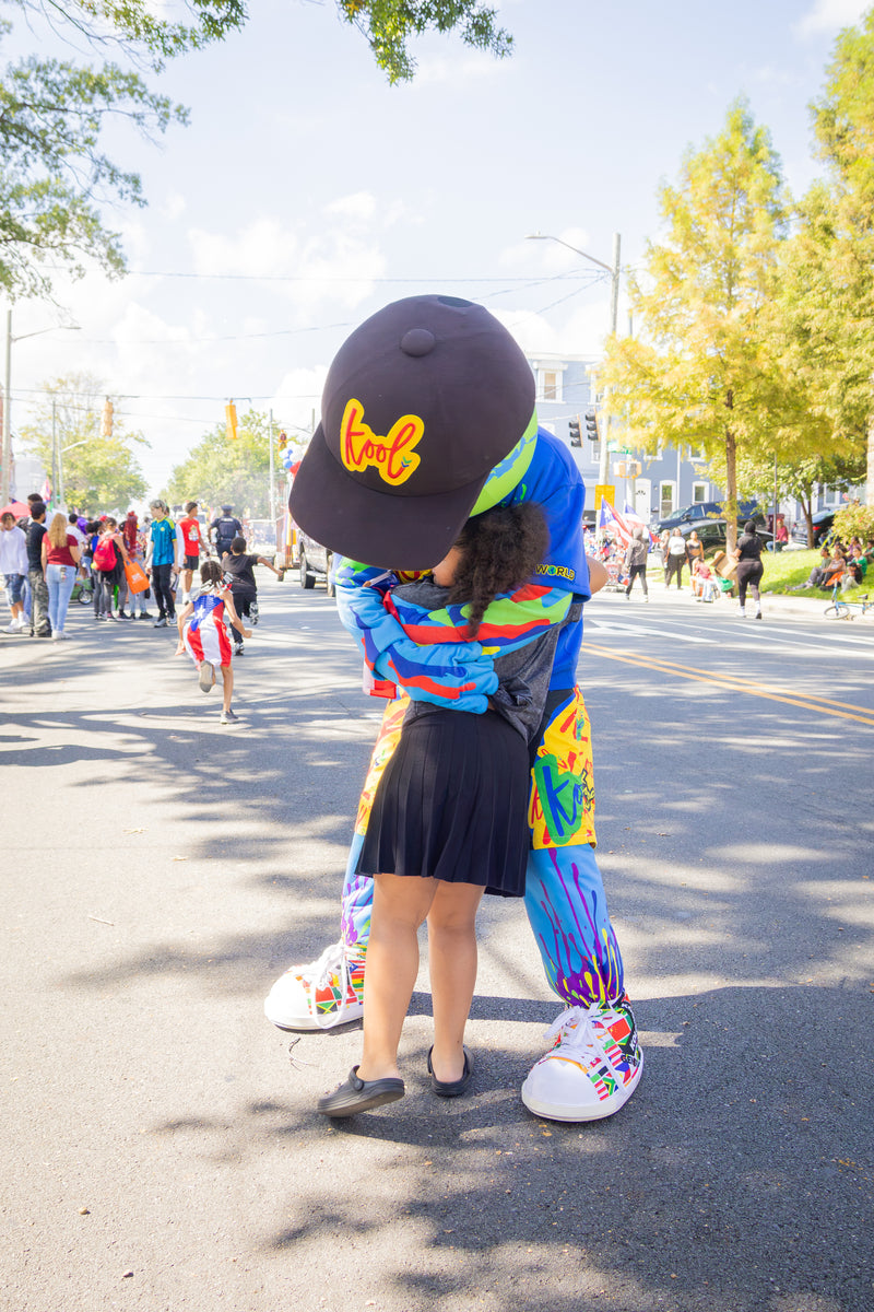 kooli in a colorful tie-dye costume and a black cap with a yellow logo kool hugs a child on a sunny outdoor street parade road.