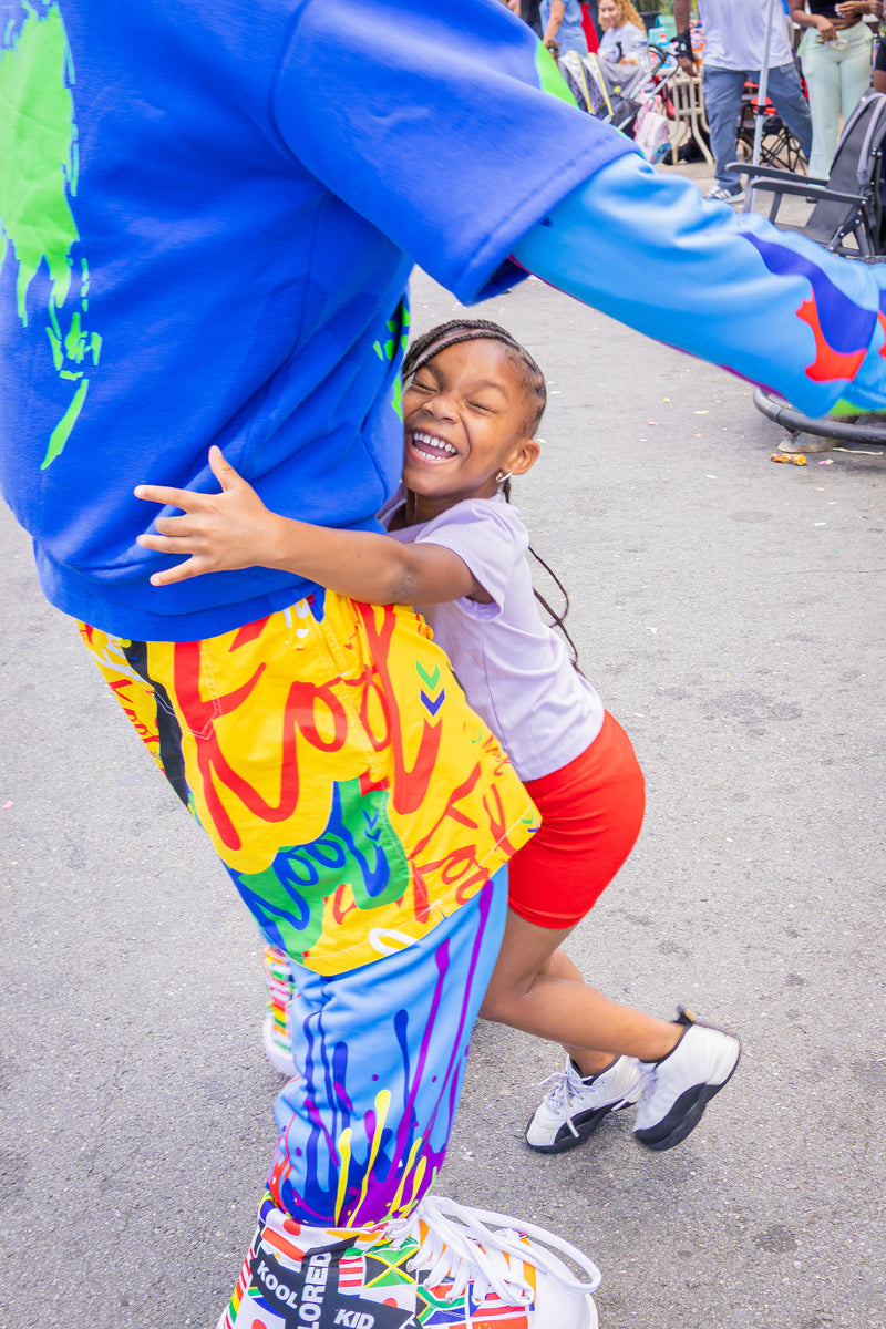 A joyful outdoor street scene featuring a child in a white t-shirt and red shorts hugging kooli wearing vibrant graffiti-style multicolor pants and a blue shirt. The child is smiling broadly, and the background shows a blurred street environment with bicycles.