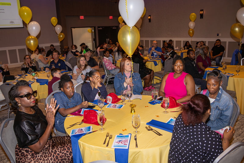 Group of people sitting at tables with yellow and blue tablecloths, clapping and smiling in a room with balloons and decorations.