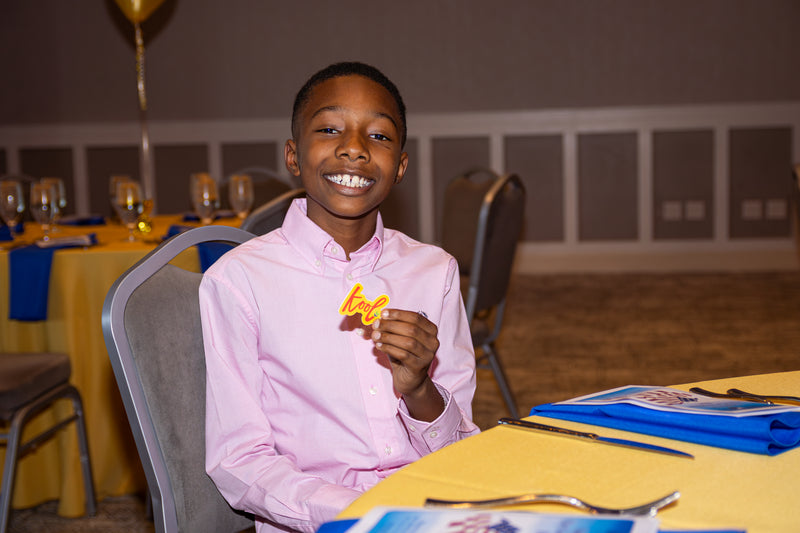 Young boy in a pink shirt holding kool sticker at a formal event with tables and chairs.