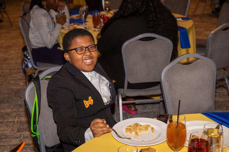Child in formal attire sitting at a table with food and drinks in a banquet hall.