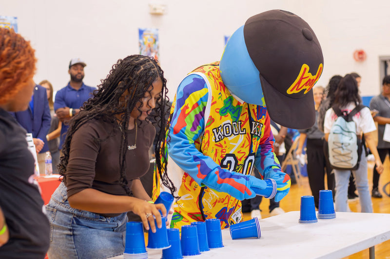 kooli pouring blue plastic cups from a table at an indoor event. A woman in a brown top and jeans is also at the table, surrounded by other people in a gymnasium.