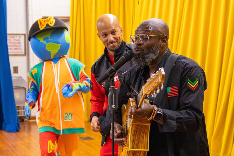 Two men, one with a microphone and the other playing guitar, standing next to a colorful mascot character against a yellow curtain.