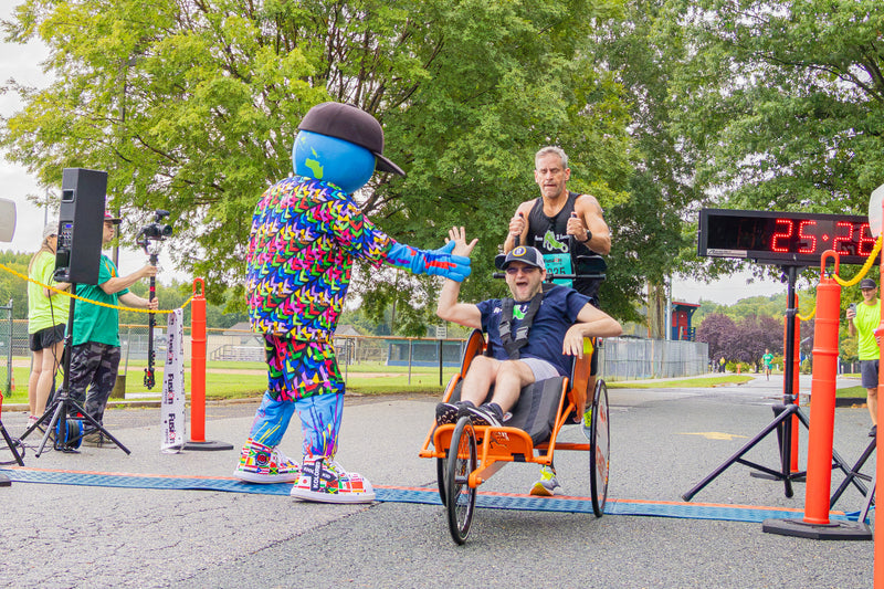 kooli waves to a man seated in an orange three-wheeled wheelchair racer at a race finish line, with a digital timer showing 25.29 and trees in the background.