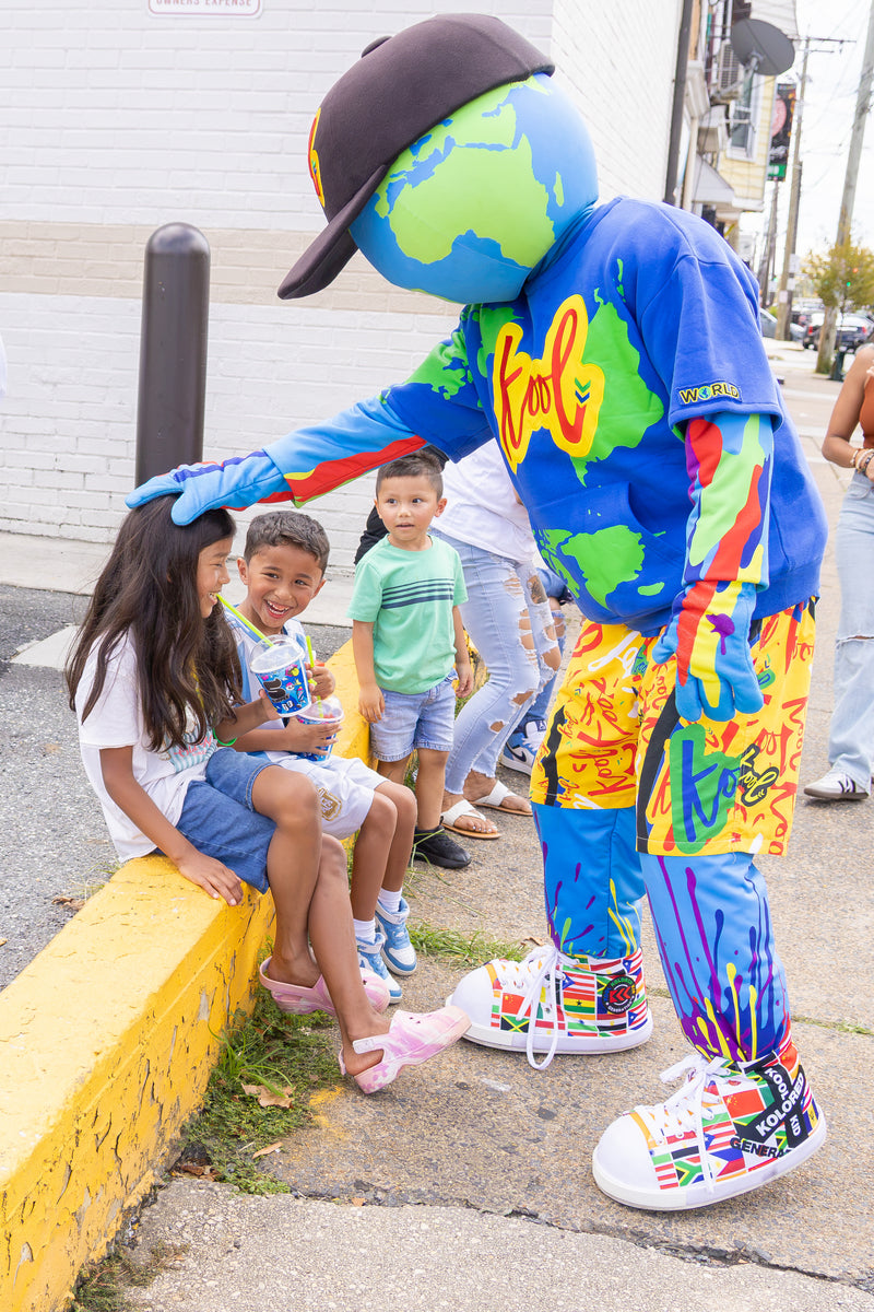 kooli stands on a sidewalk next to three children sitting on a yellow curb.