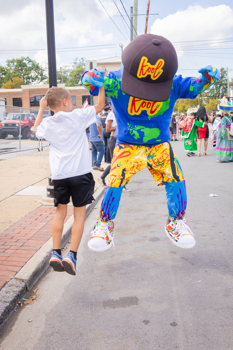 A child in a white t-shirt and black shorts jumps in the air holding the hands of kooli wearing a dark brown cap with yellow 'Kool' lettering, a blue shirt, and bright multicolor shorts, set on a street during a parade.