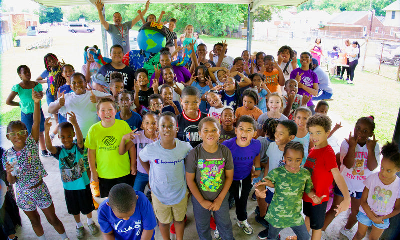 A large group of children and adults posing together under a white canopy outdoors, with kooli visible in the center. 