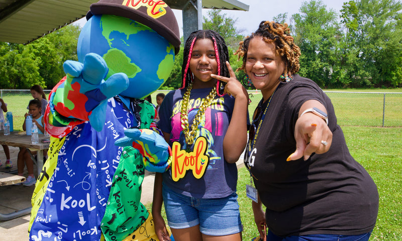 Two women and Kooli pose together outdoors at a park  under a canopy. The woman on the right points toward the camera, wearing a black shirt and blue jeans.