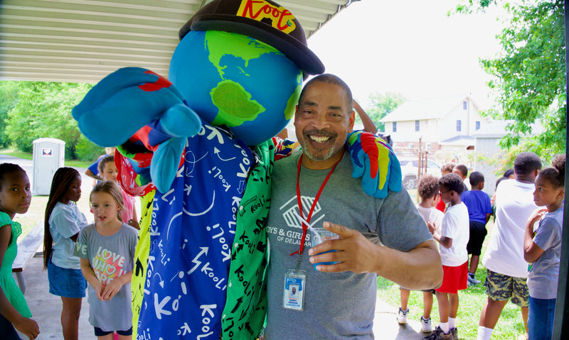 A smiling man in a gray t-shirt stands outdoors under a sheltered porch next to a large blue and green globe mascot wearing a black cap with 'Kooli' Children are visible in the background.