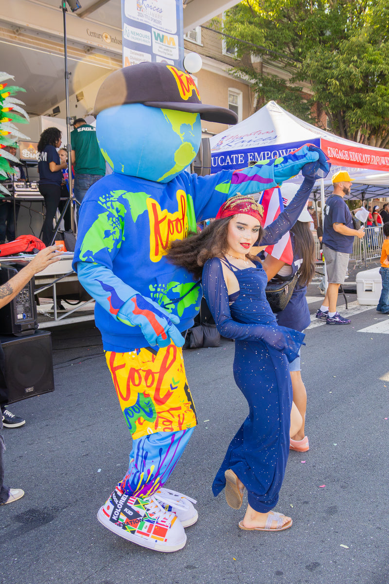 kooli dances outdoors with a woman in a blue dress at a street festival. Tents, stage equipment, and other people are visible in the background.
