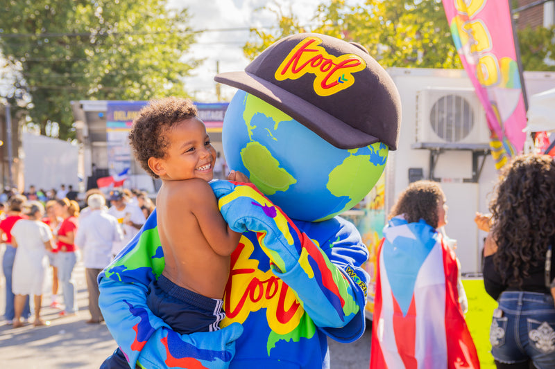 kooli holding a toddler, with a crowd and outdoor event background visible.